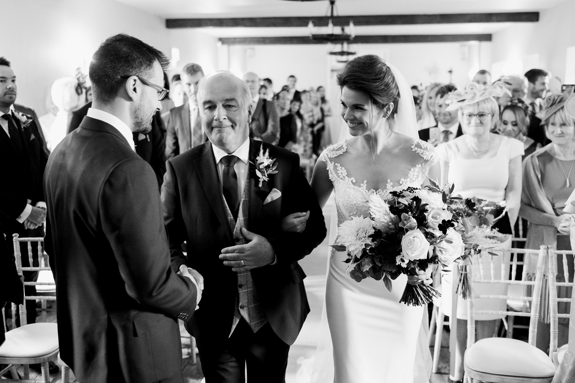 father of bride shakes grooms hand at top of aisle as bride smiles on